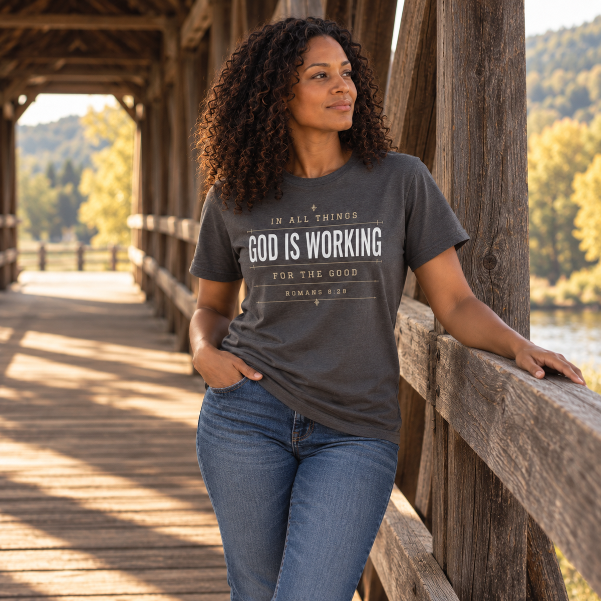 Woman wearing the graphite Romans 8:28 “God Is Working” Christian shirt while leaning naturally against the railing of a historic wooden covered bridge.