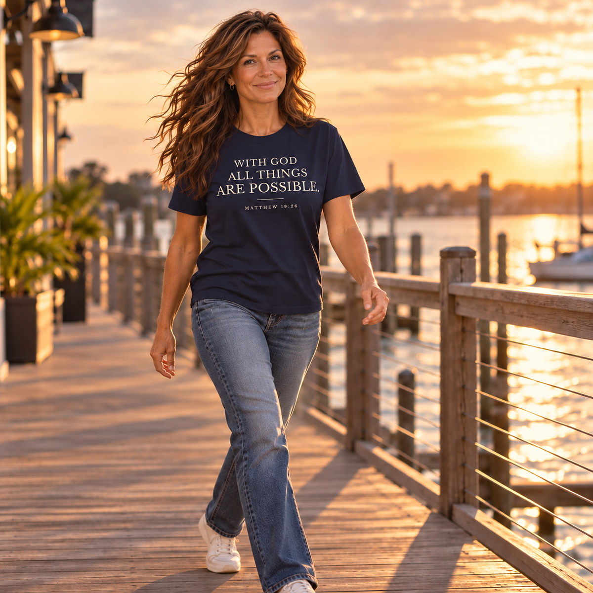 Woman walking along a wooden waterfront pier wearing a navy With God All Things Are Possible T-shirt.