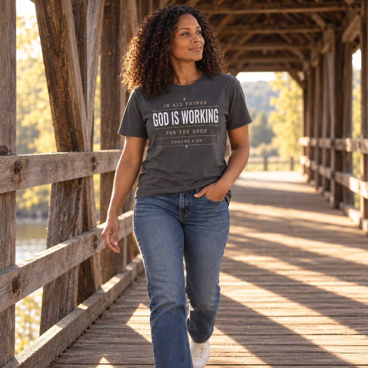Woman walking through a sunlit covered bridge wearing a graphite “God Is Working for the Good” Romans 8:28 Christian T-shirt with blue jeans.