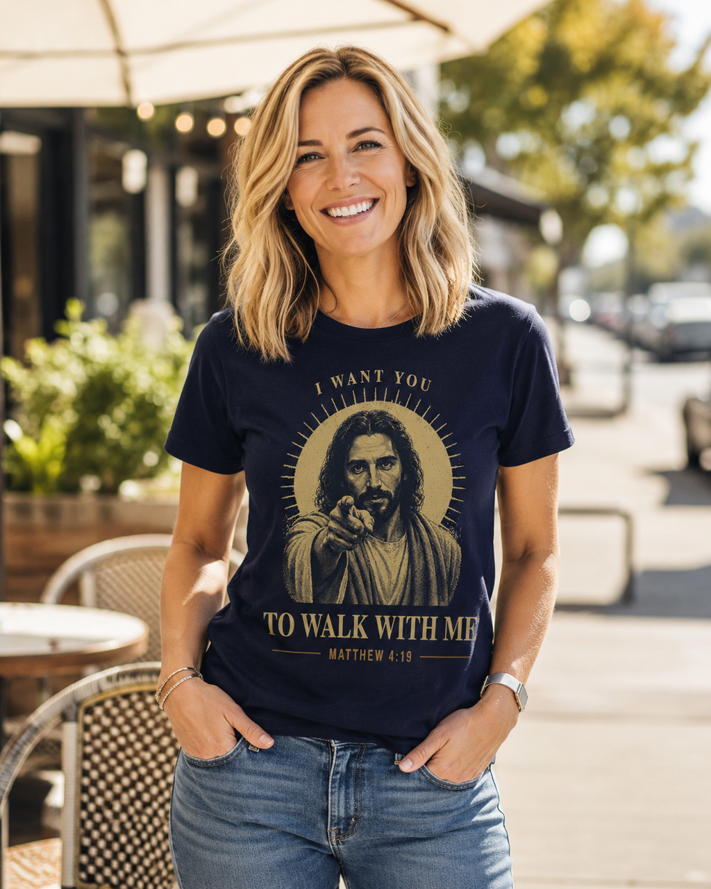 A smiling blonde woman stands outdoors wearing a navy graphic T-shirt with a large beige illustration of Jesus and the text 'I WANT YOU TO WALK WITH ME MATTHEW 4:19.