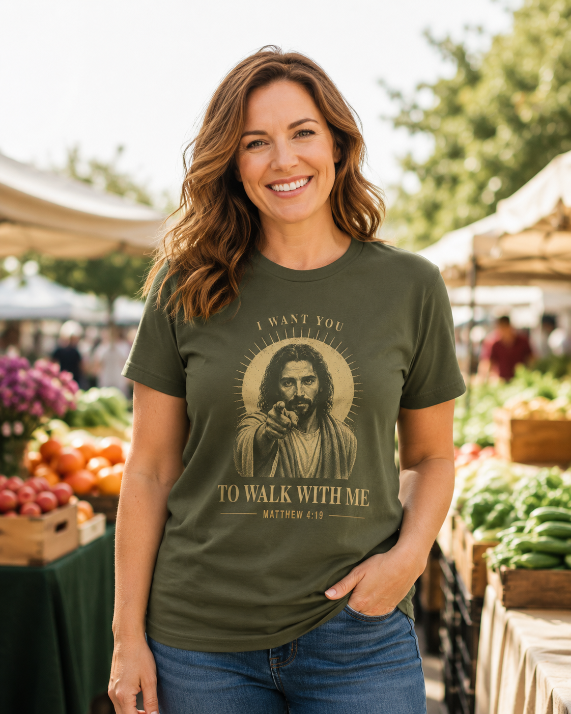 Smiling woman wearing an olive green Christian T-shirt featuring Jesus and the words “I Want You to Walk With Me,” inspired by Matthew 4:19, at an outdoor market.