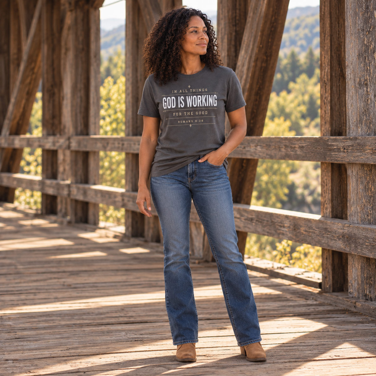 Full-body view of woman wearing a graphite “God Is Working” Romans 8:28 Christian T-shirt with blue jeans and casual boots inside a covered bridge.