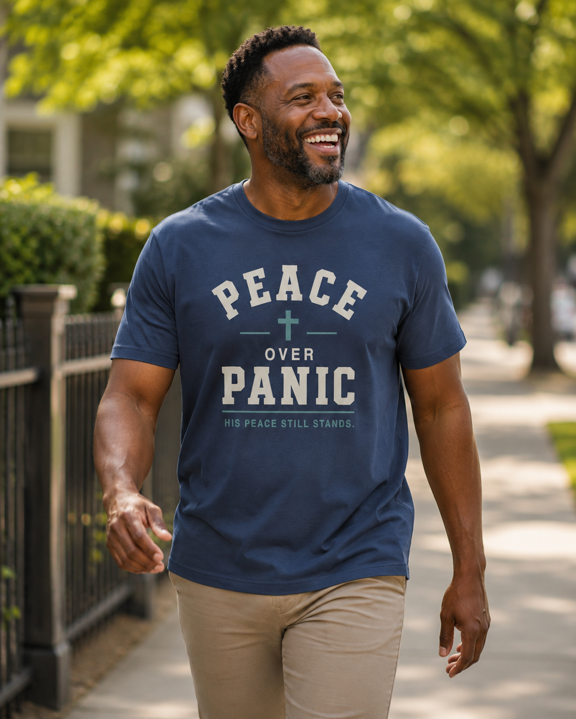 A man walking outdoors on a sunny sidewalk wearing a navy blue graphic t-shirt with the text 'PEACE OVER PANIC' and 'HIS PEACE STILL STANDS.' in white and light green, paired with khaki pants.