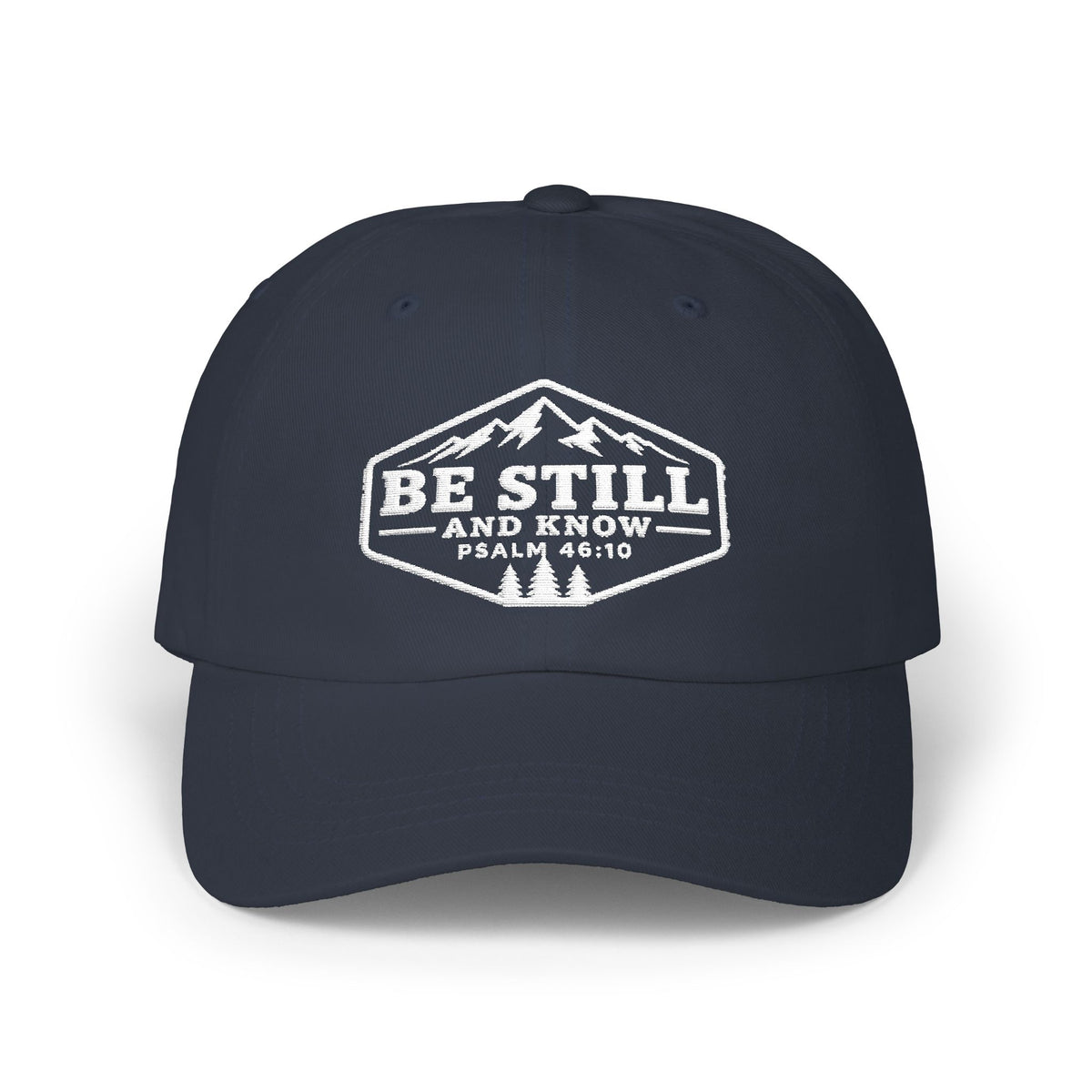 A navy blue baseball cap with a white embroidered graphic on the front that reads 'BE STILL AND KNOW PSALM 46:10' with a mountain and pine tree design, shown on a clean white background.