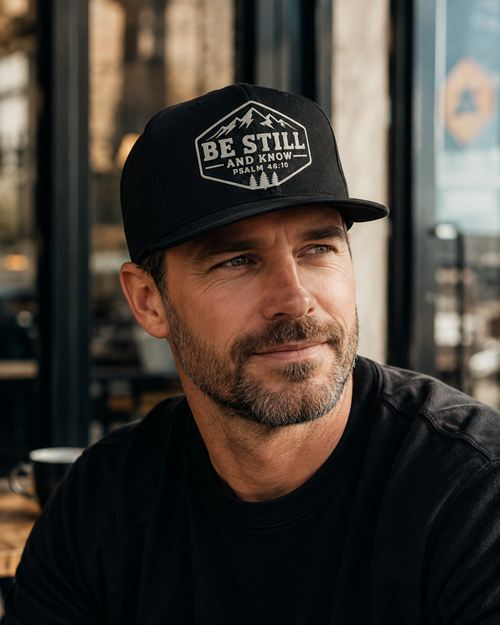 Man wearing black Be Still and Know Psalm 46:10 Christian hat with embroidered mountain and pine design.