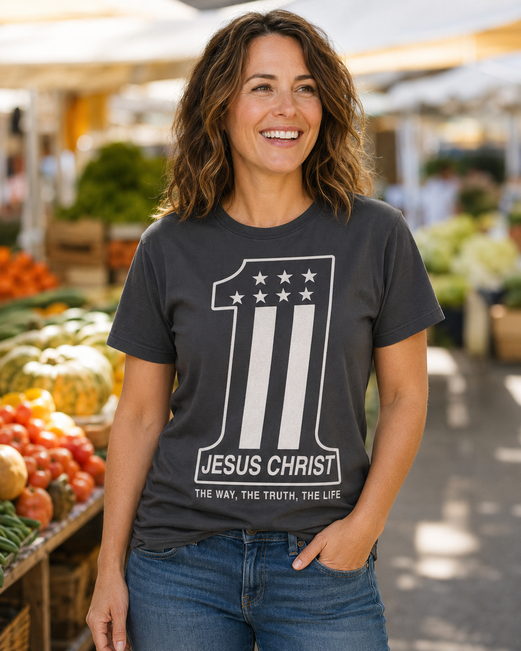 A smiling woman wearing a dark charcoal gray graphic t-shirt with a white '1' design featuring stars and stripes, and the text 'JESUS CHRIST' and 'THE WAY, THE TRUTH, THE LIFE'. She is standing in a blurred outdoor market setting with produce.
