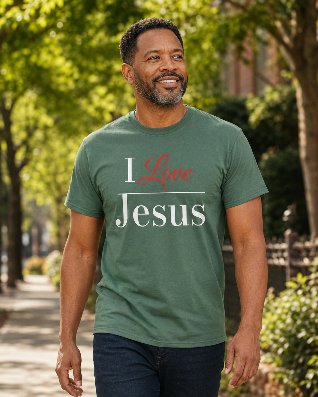 A man wearing a sage green graphic t-shirt with the text 'I Love Jesus' printed on the front, walking outdoors on a tree-lined path.