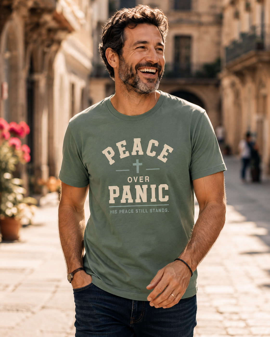 Smiling man wearing a sage green “Peace Over Panic” Christian T-shirt while walking along a sunlit city street.