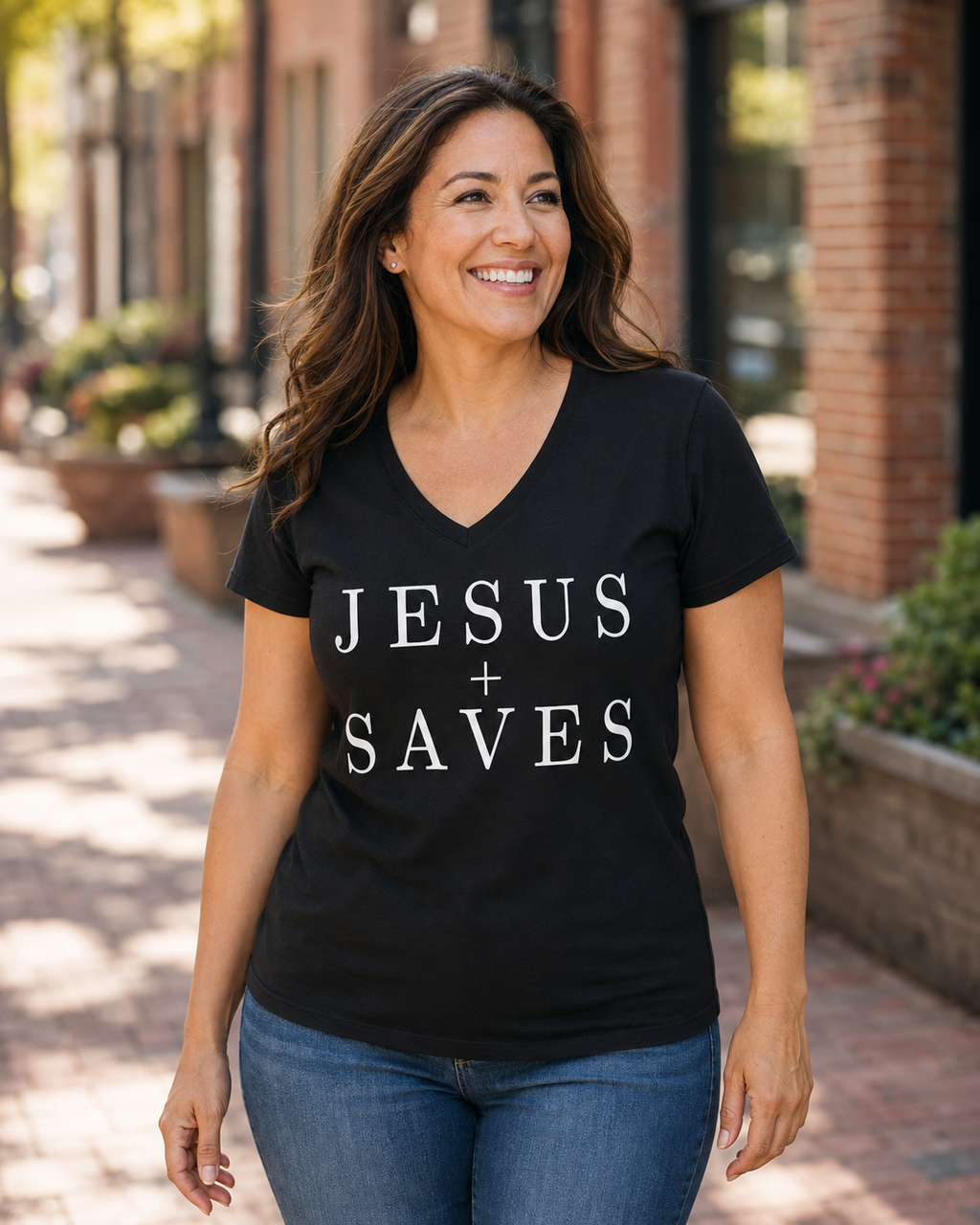 A smiling woman wearing a black V-neck t-shirt with the text 'JESUS + SAVES' printed in white capital letters, standing outdoors on a brick sidewalk.