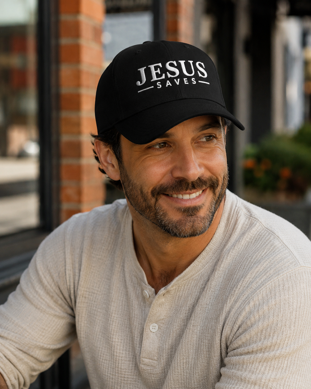 A smiling man wearing a black baseball cap with white embroidered text reading 'JESUS SAVES' and a light beige henley shirt, photographed outdoors in front of a brick building.