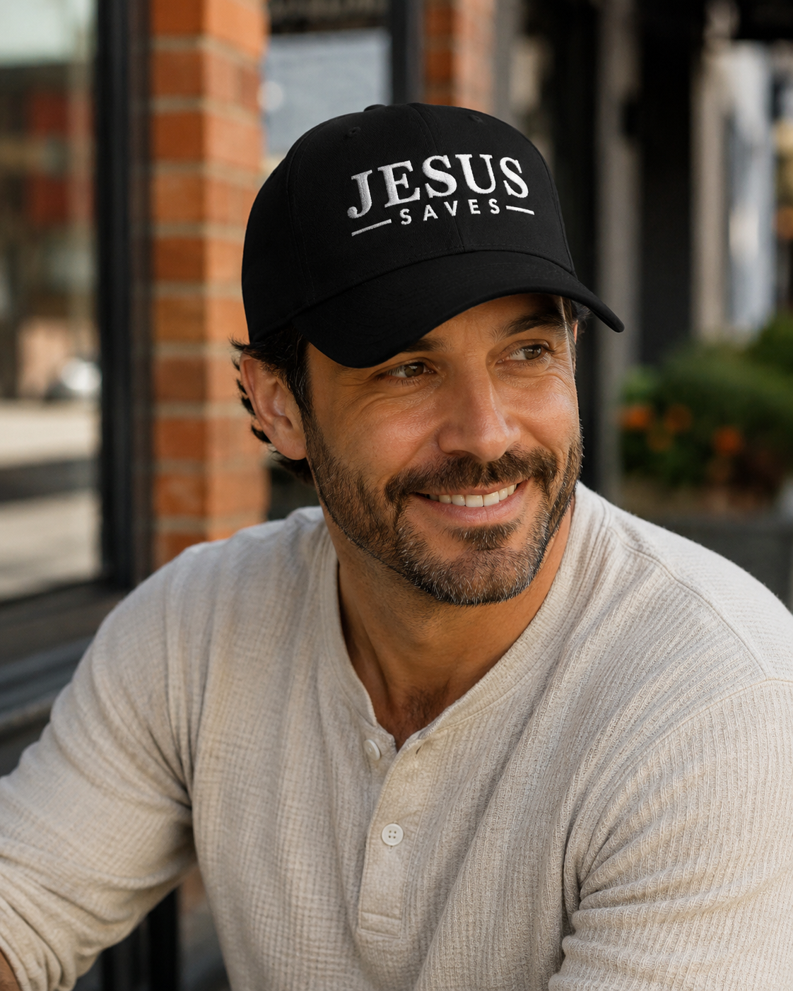 A smiling man wearing a black baseball cap with white embroidered text reading 'JESUS SAVES' and a light beige henley shirt, photographed outdoors in front of a brick building.