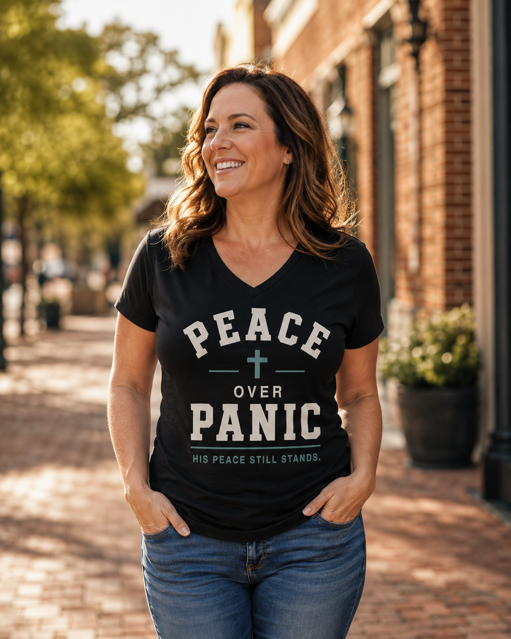 A woman wearing a black V-neck graphic t-shirt with the text 'PEACE OVER PANIC' and 'HIS PEACE STILL STANDS.' walking down a sunny brick street.