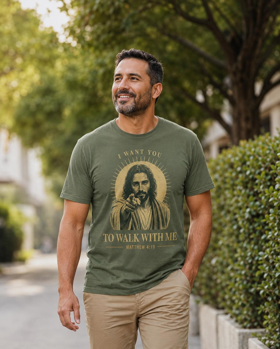 A man wearing an olive green graphic t-shirt with a vintage-style print of Jesus and the text 'I WANT YOU TO WALK WITH ME - MATTHEW 4:19 walking outdoors on a tree-lined path.