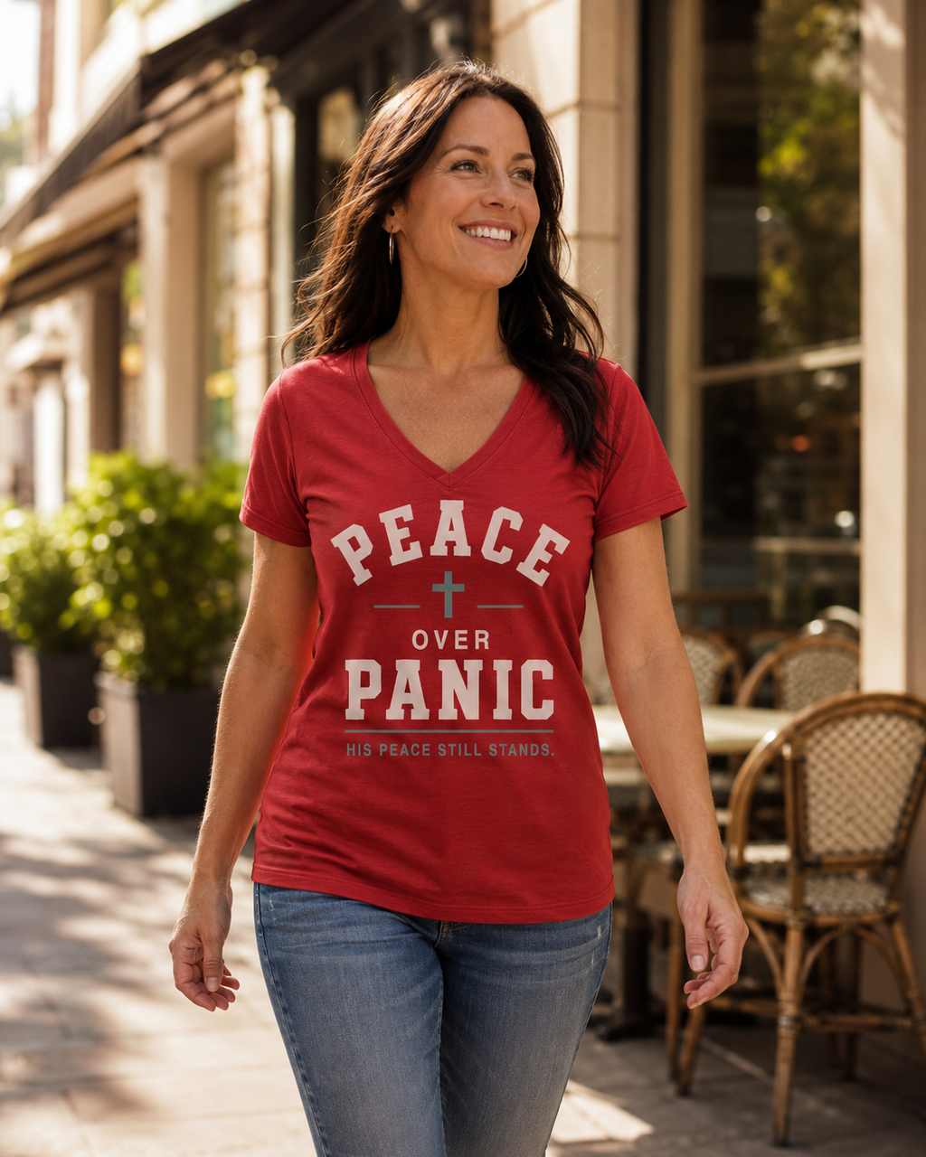 A woman walking outdoors on a city sidewalk wearing a red V-neck graphic T-shirt with the words 'PEACE OVER PANIC' and smaller text 'HIS PEACE STILL STANDS.' She is also wearing blue jeans, with storefronts, plants, and outdoor chairs in the background.