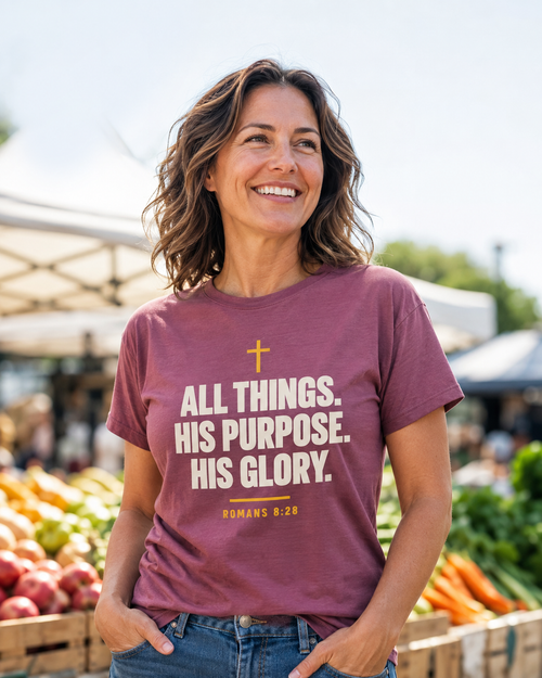 Woman wearing a berry Christian T-shirt featuring “All Things. His Purpose. His Glory.” with a cross and Romans 8:28 artwork outdoors.