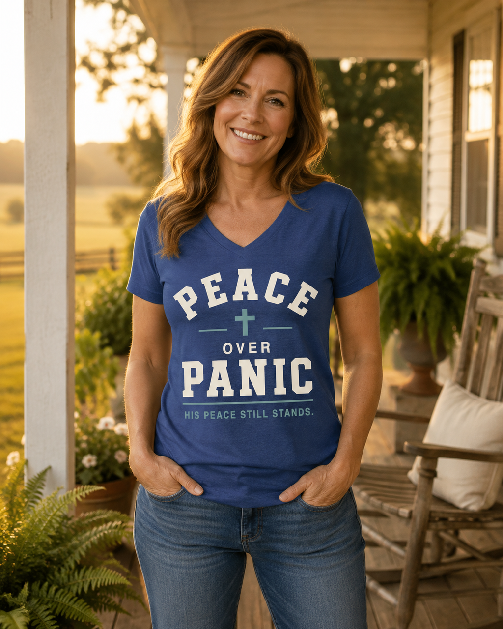 A smiling woman stands on a porch wearing a blue V-neck T-shirt with the words “PEACE OVER PANIC” and smaller text “HIS PEACE STILL STANDS.” She is also wearing blue jeans, with a warm outdoor sunset background and porch furniture visible.