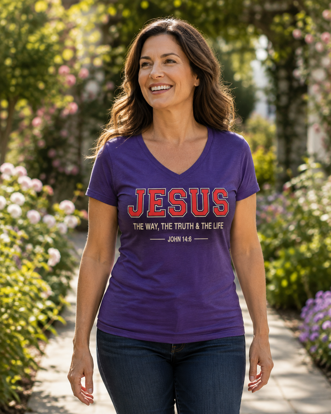 A smiling woman wearing a purple V-neck t-shirt with 'JESUS THE WAY, THE TRUTH & THE LIFE' and 'JOHN 14:6' printed on the front, walking outdoors on a garden path.