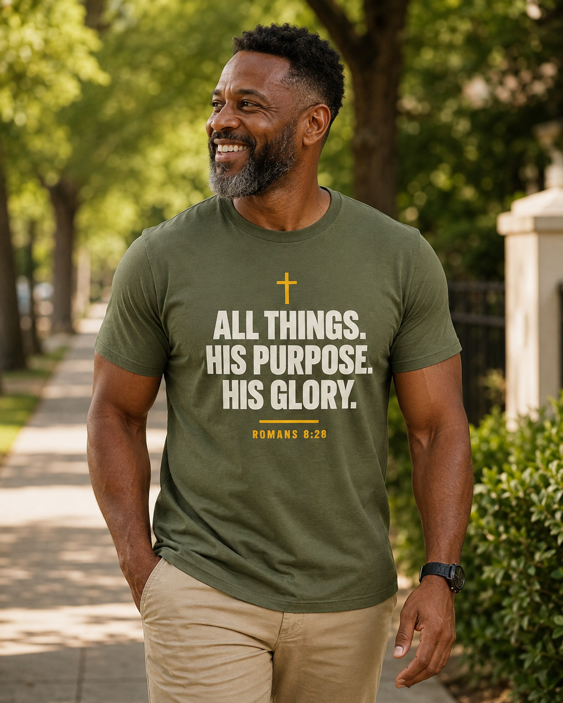Smiling man wearing an olive green Christian T-shirt reading “All Things. His Purpose. His Glory.” with Romans 8:28 while walking outdoors.