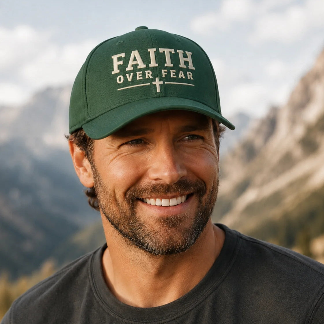 A smiling man outdoors wearing a dark green baseball cap with the words 'FAITH OVER FEAR' and a small cross emblem on the front, paired with a dark gray t-shirt against a blurred mountain background.