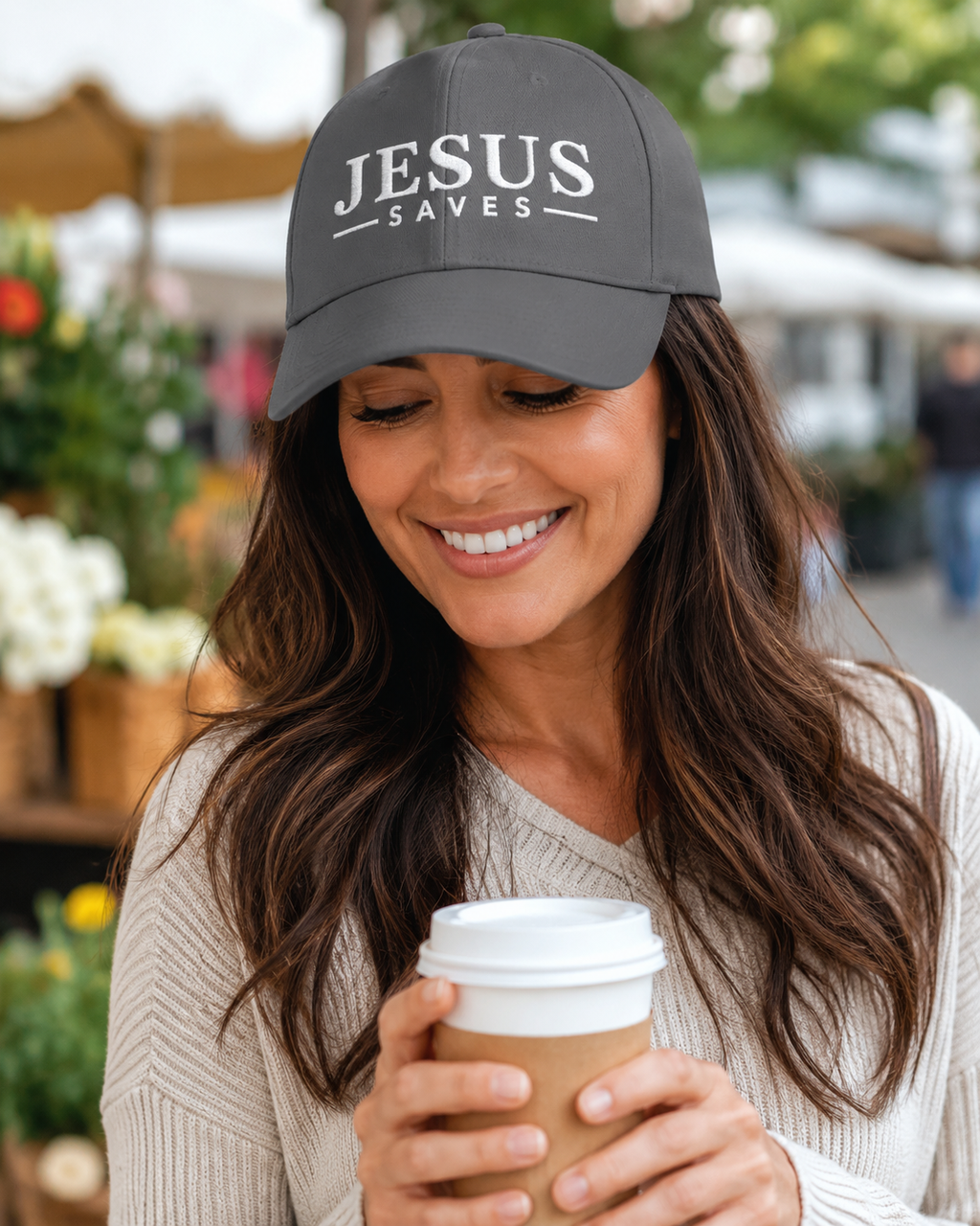 A smiling woman wearing a dark gray baseball cap with the white text 'JESUS SAVES' holds a paper coffee cup in an outdoor market setting.