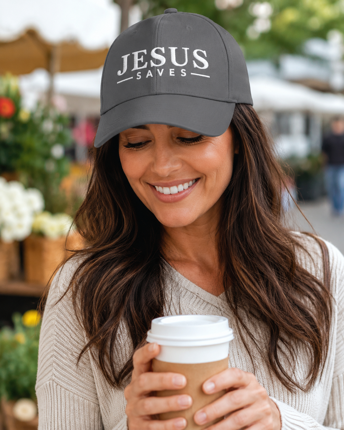 A smiling woman wearing a dark gray baseball cap with the white text 'JESUS SAVES' holds a paper coffee cup in an outdoor market setting.