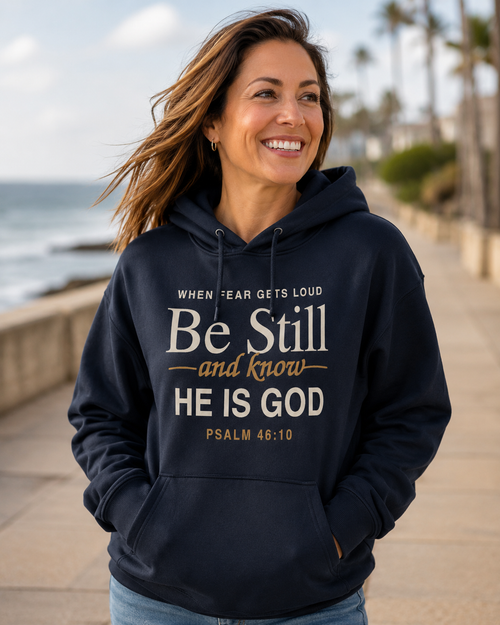 A woman smiling outdoors on a beach boardwalk wearing a navy hoodie with a large white and gold Bible verse graphic on the front: 'When fear gets loud Be Still and know He is God Psalm 46:10'.