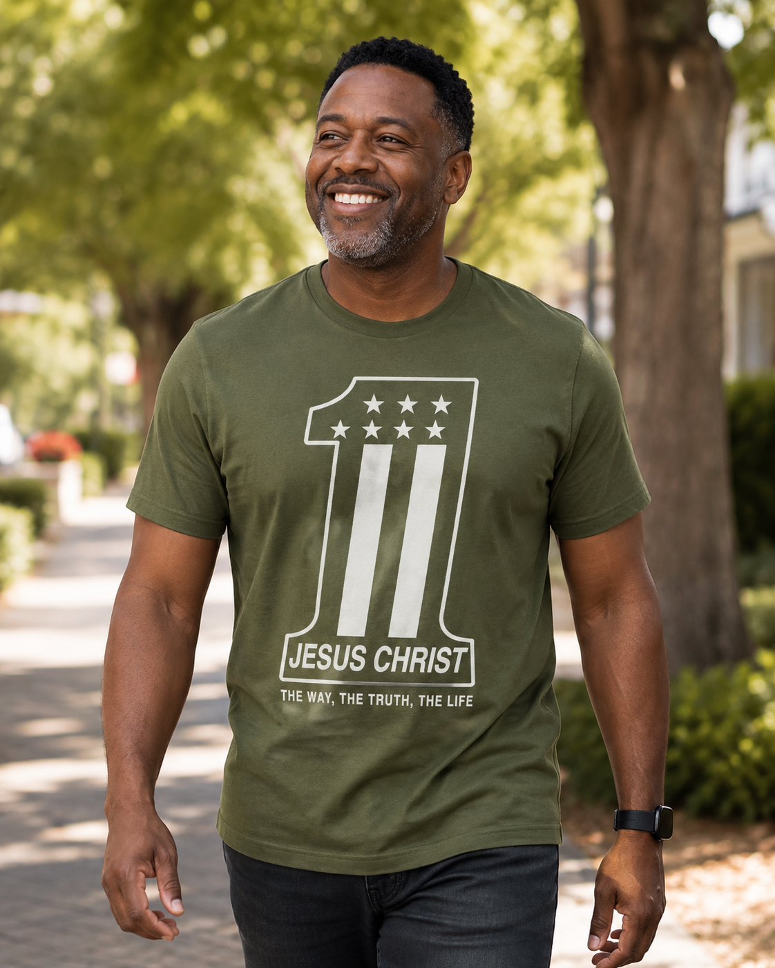 A smiling man wearing an olive green t-shirt with a white graphic print that reads '1 JESUS CHRIST' and 'THE WAY, THE TRUTH, THE LIFE' walking outdoors on a tree-lined path.