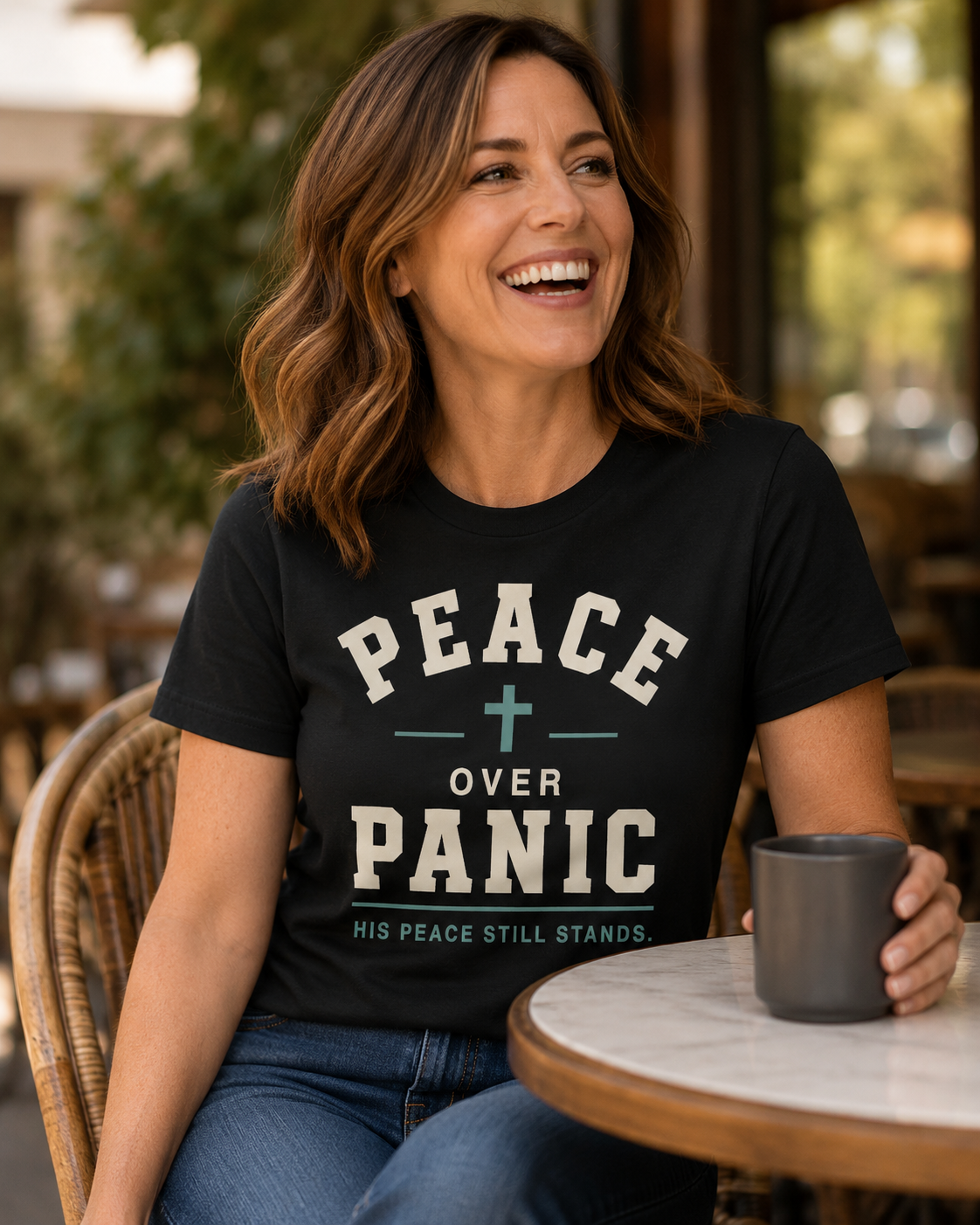 Smiling woman wearing a black “Peace Over Panic” Christian T-shirt while holding a mug at an outdoor café.