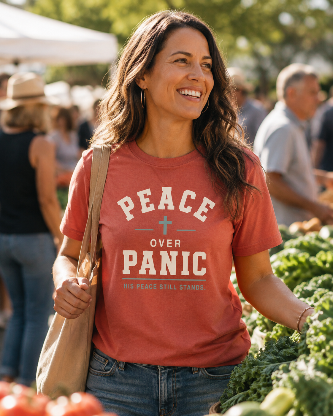 A woman smiling at a farmers market wearing a rust-orange graphic t-shirt that reads 'PEACE OVER PANIC' with a small cross and the phrase 'HIS PEACE STILL STANDS.' below it.