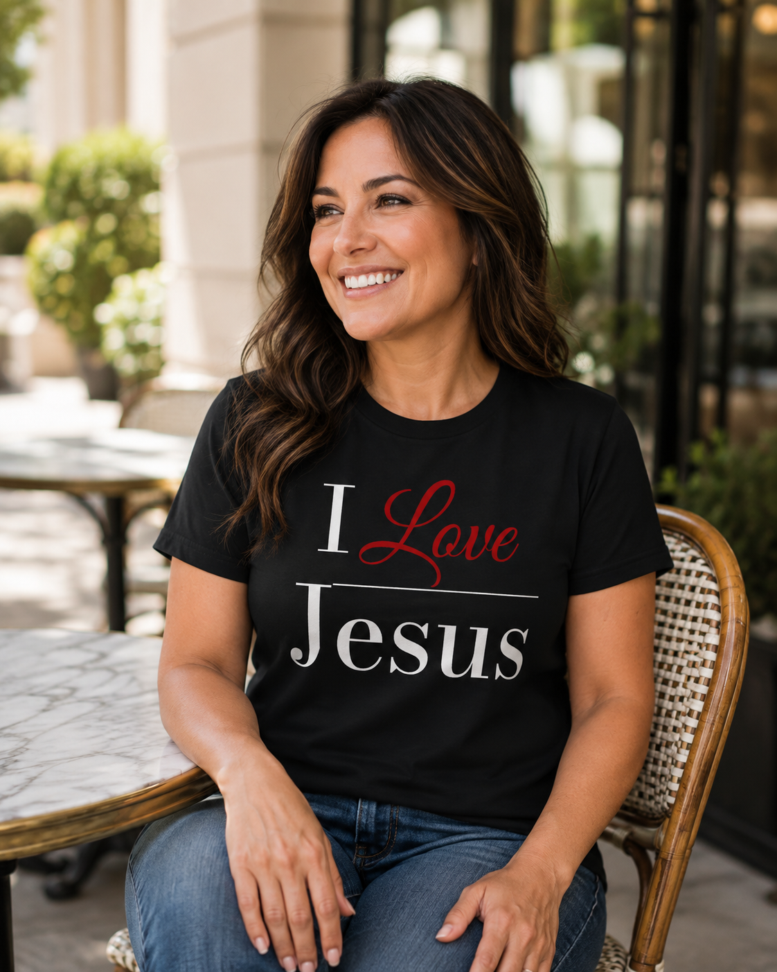 A smiling woman with long brown hair sits on a woven-back chair at an outdoor cafe, wearing a black graphic t-shirt that reads 'I Love Jesus' in white and red lettering, paired with blue jeans.