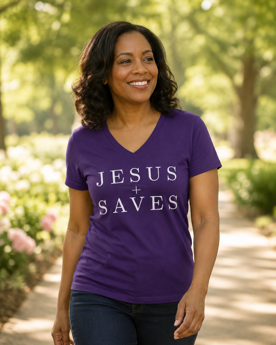 A woman wearing a purple V-neck t-shirt with the text 'JESUS + SAVES' printed in white, standing in a sunny park with trees and a path in the background.