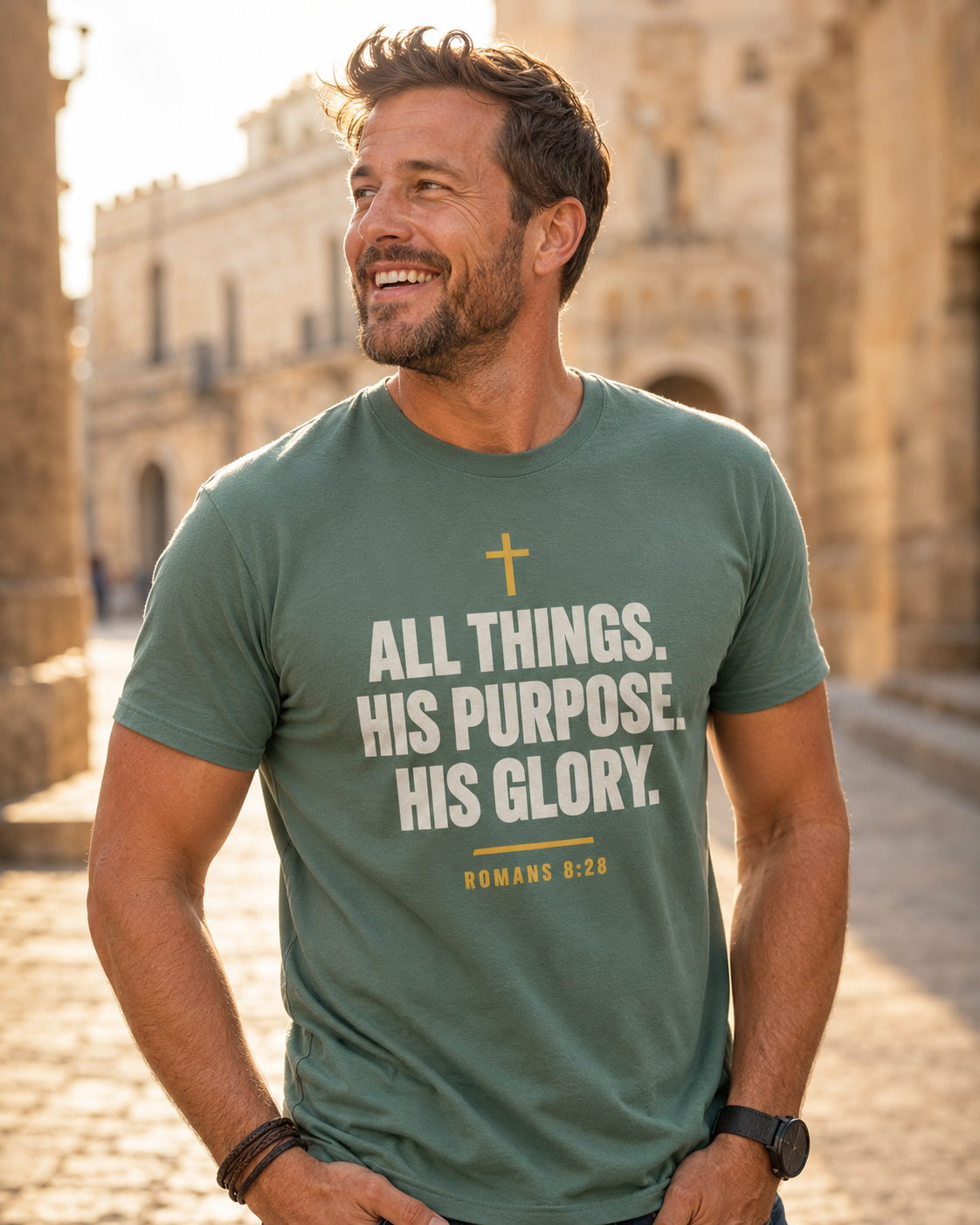 Man wearing a sage green Christian T-shirt featuring “All Things. His Purpose. His Glory.” with a cross and Romans 8:28 outdoors near a stone building.