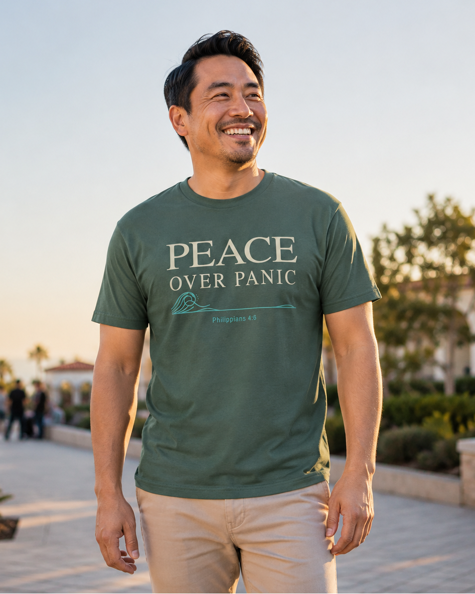 A smiling man wearing an olive green graphic t-shirt with the text 'PEACE OVER PANIC' and khaki pants, standing outdoors in a sunny, paved plaza with blurred background elements.